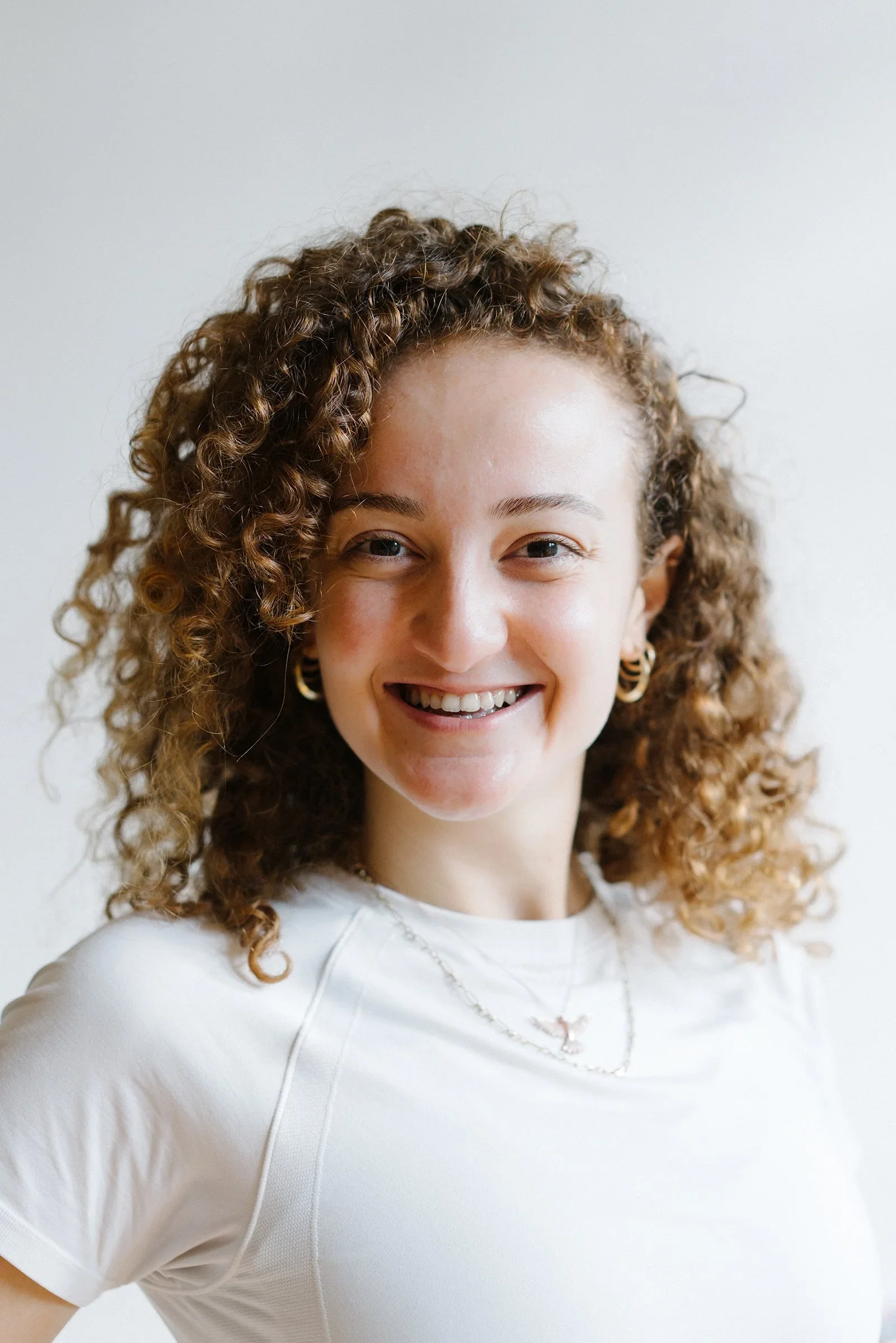 Close-up of a young woman with curly hair smiling, wearing hoop earrings, a necklace, and a white shirt, standing against a light background.