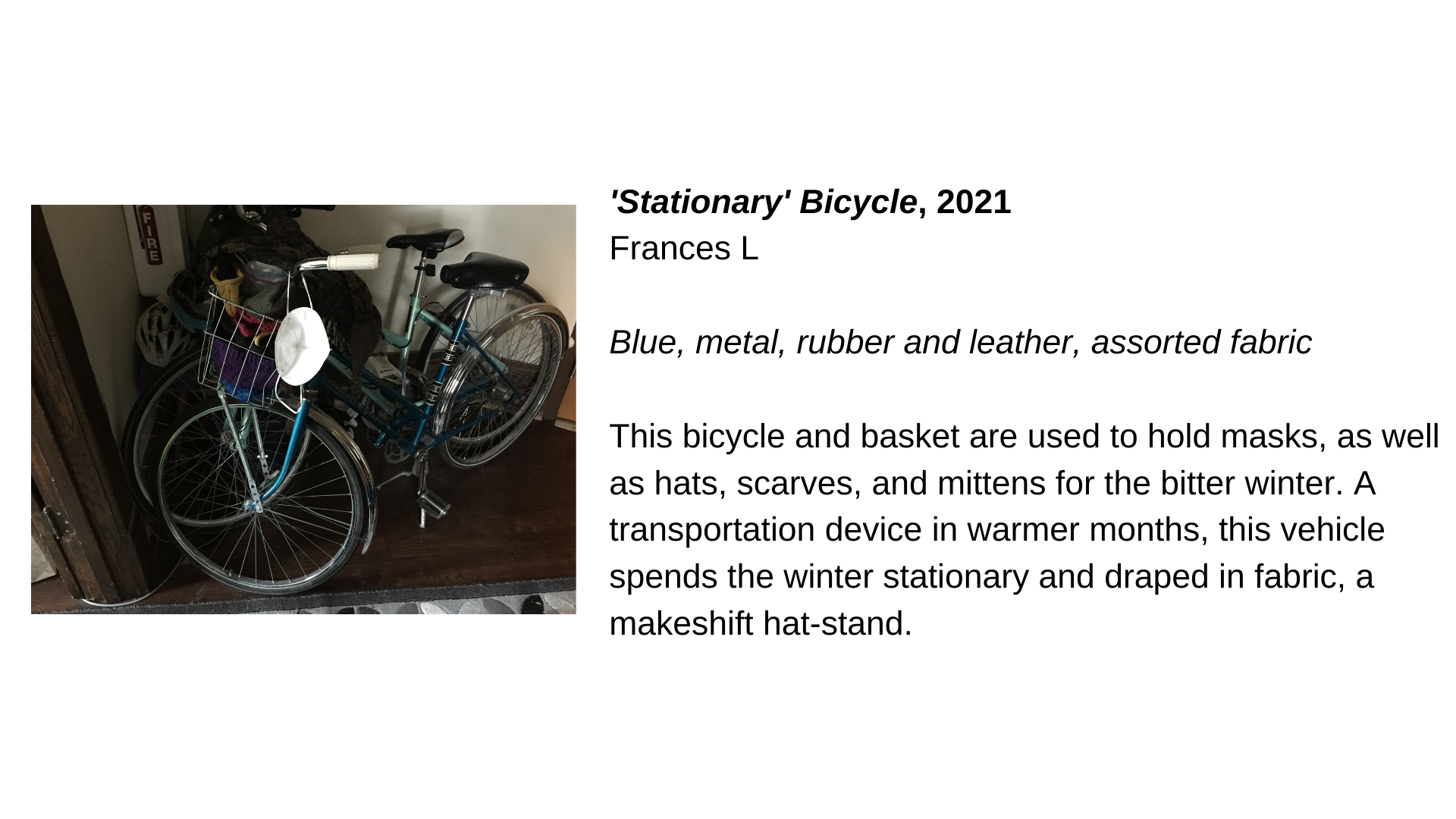  A photograph of a bicycle next to the text, “'Stationary' Bicycle, 2021 - Frances L. Blue, metal, rubber and leather, assorted fabric. This bicycle and basket are used to hold masks, as well as hats, scarves, and mittens for the bitter winter. A tra