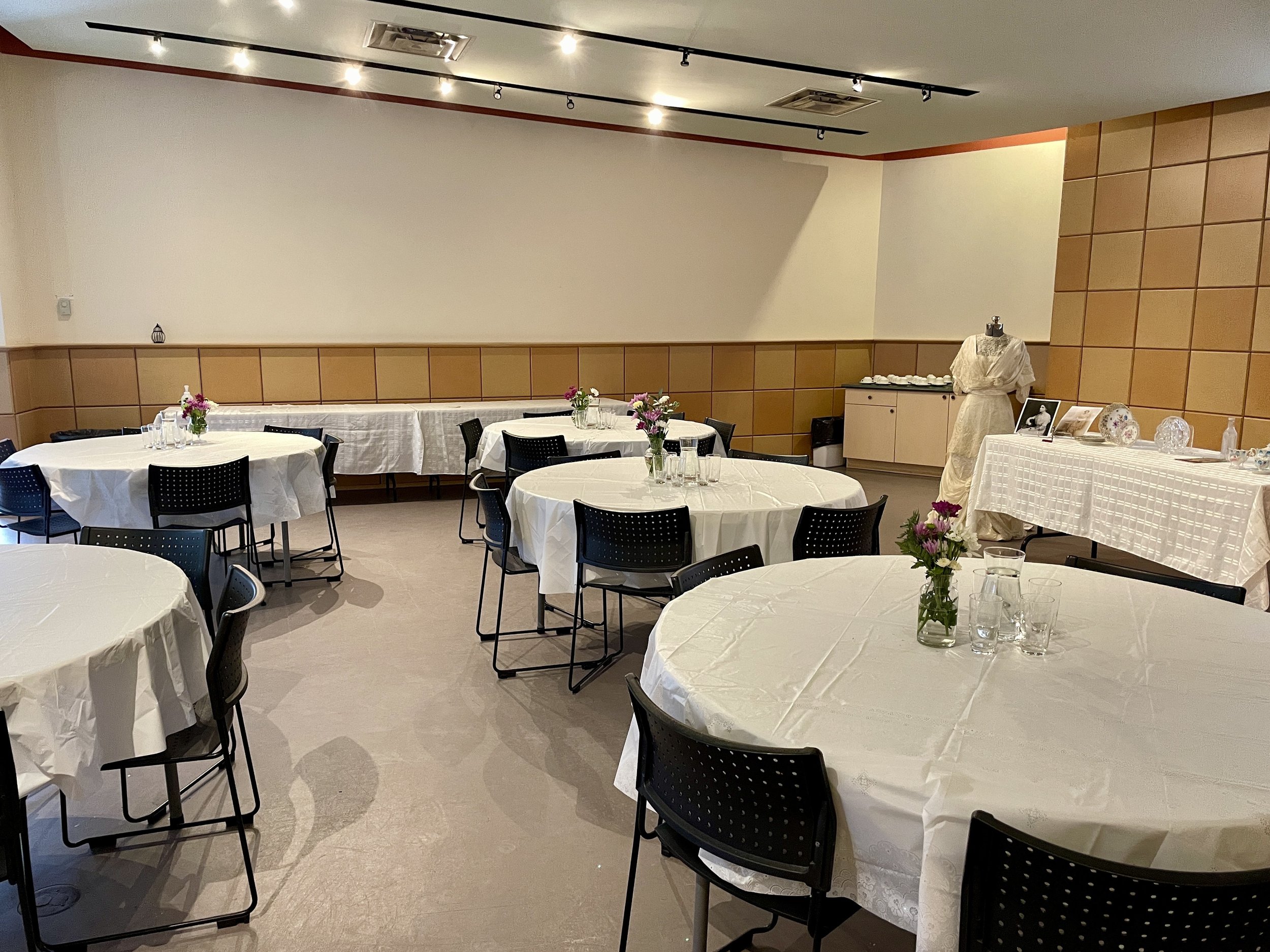  Circular tables with white tablecloths and floral centerpieces in the Visitors’ Centre. A dress form with a wedding dress on it sits at the top of the room alongside a rectangular displaying artifacts.  