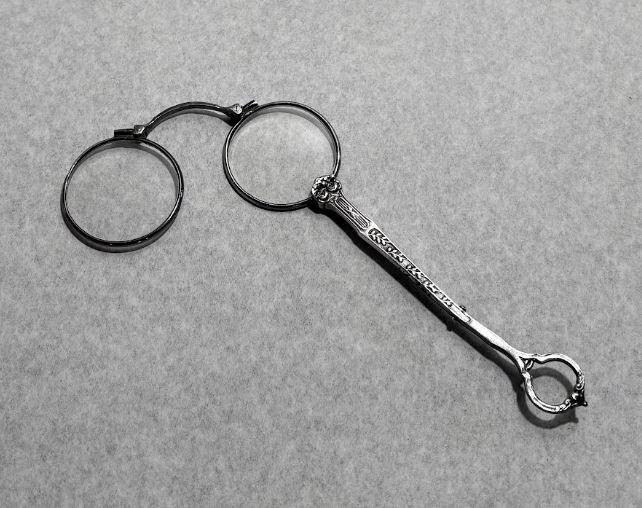 a hand-held pair of eye glasses with circular lenses. The silver handle is delicate, with floral engravings and a ring at the end.