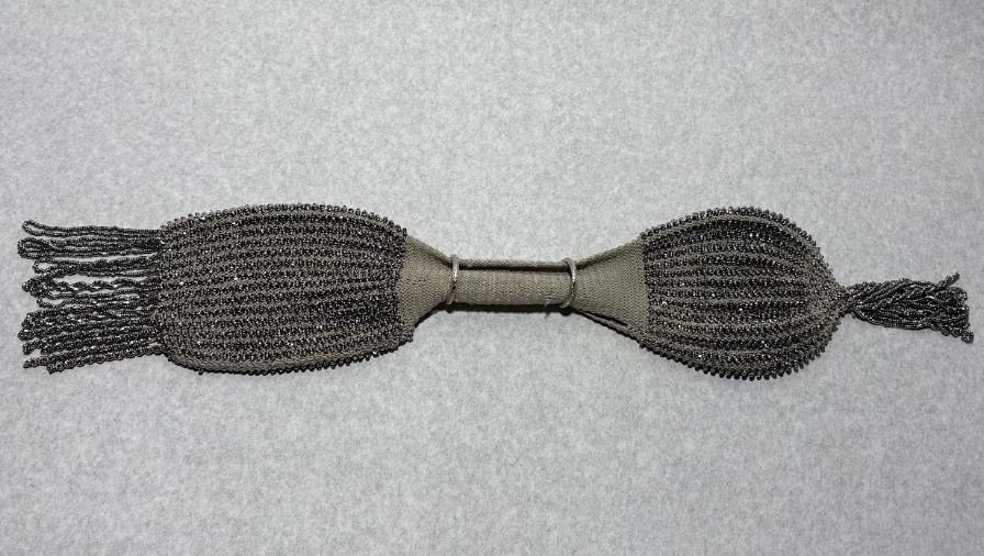 a grey crocheted change purse with silver beading. The purse is shaped like an hourglass, and has 2 silver rings near the centre. The ends of the purse have beaded tassels.