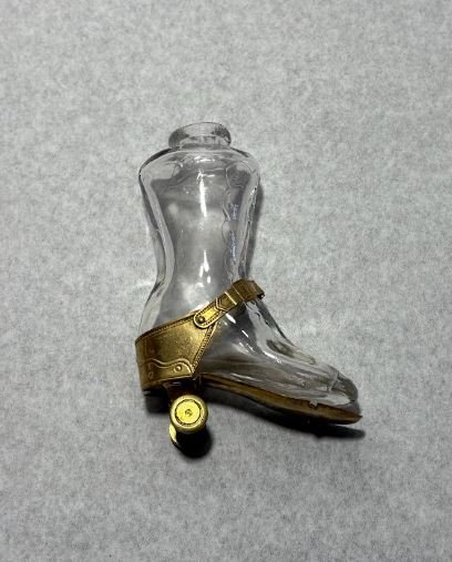 A pressed glass perfume bottle in the shape of a boot. It has a gold-coloured brass heel and footplate, with two back wheels (that actually roll). The two front wheels are missing.