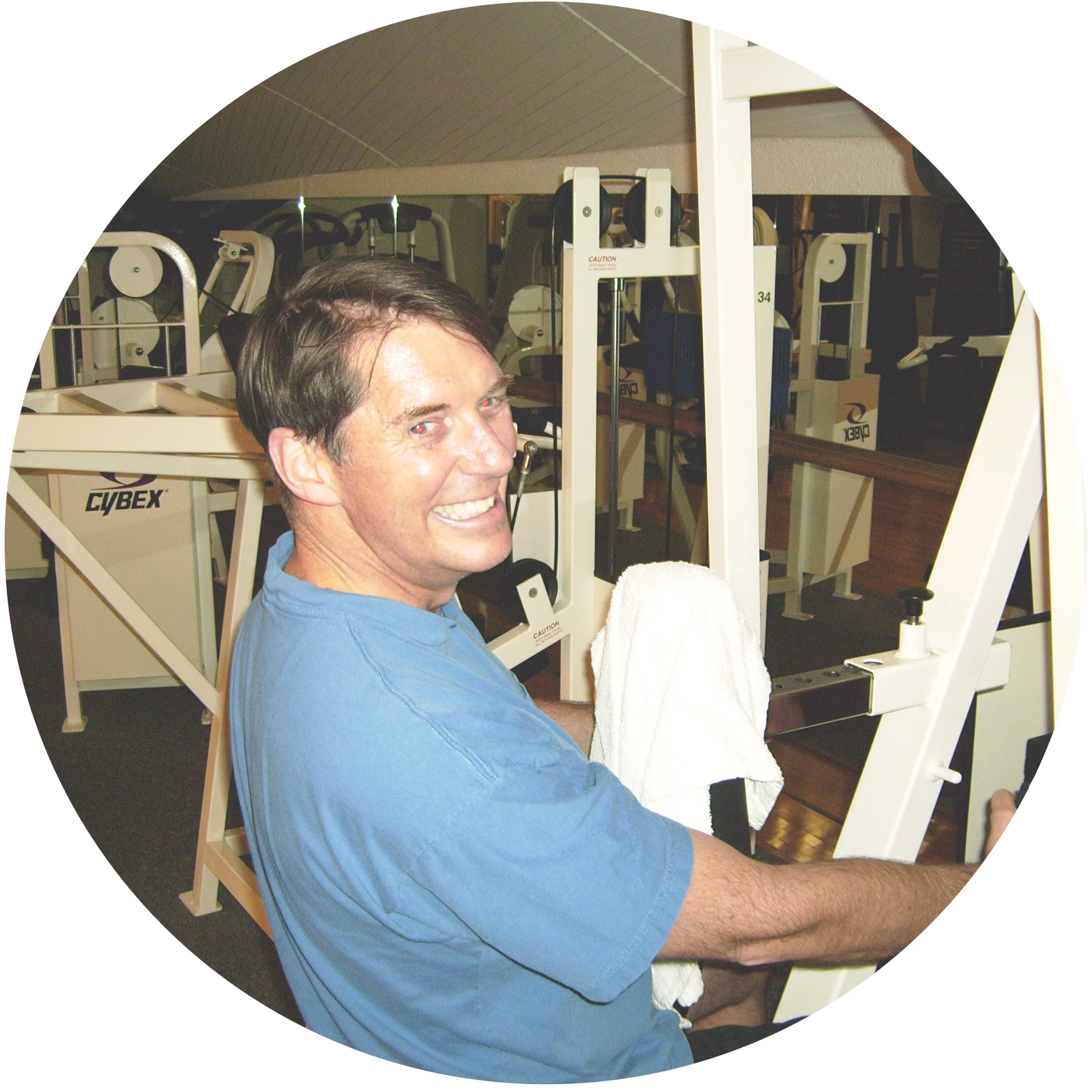 A man with dark hair smiling while sitting on gym equipment in a fitness room.