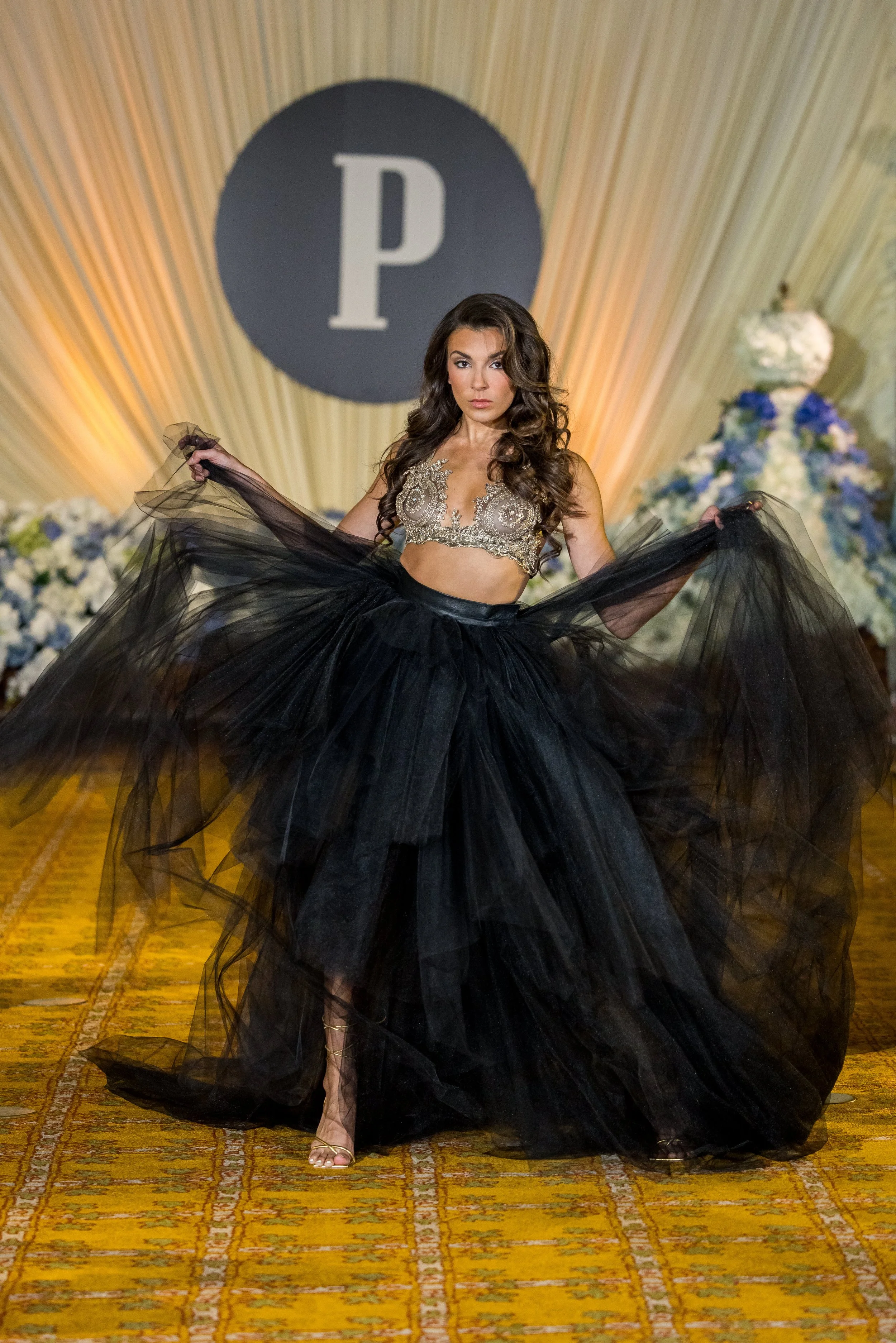 A woman in a gold and silver embroidered crop top and a full black tulle ball gown, posing on a runway with a large letter 'P' in the background and floral arrangements on either side.
