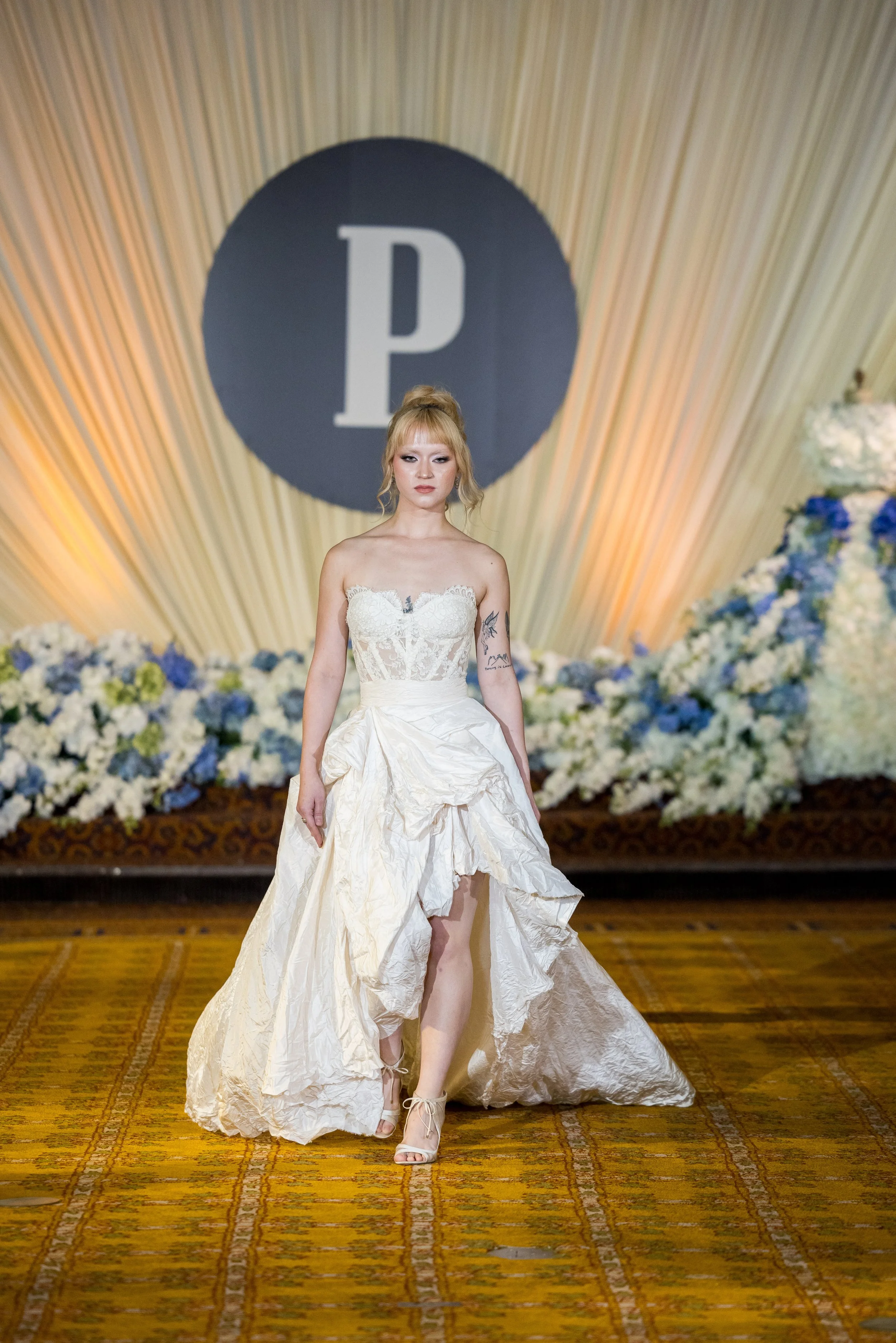A woman with blonde hair in an updo wearing a strapless white wedding gown with a ruffled skirt is walking on a patterned yellow and red carpet. Behind her is a large blue circle with a white letter 'P' and a backdrop of flowing drapes with a floral 
