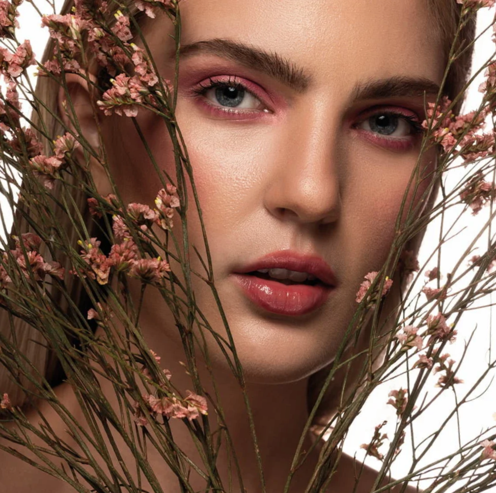 A close-up of a woman's face surrounded by pink flowers and greenery, with detailed makeup and glossy lips.