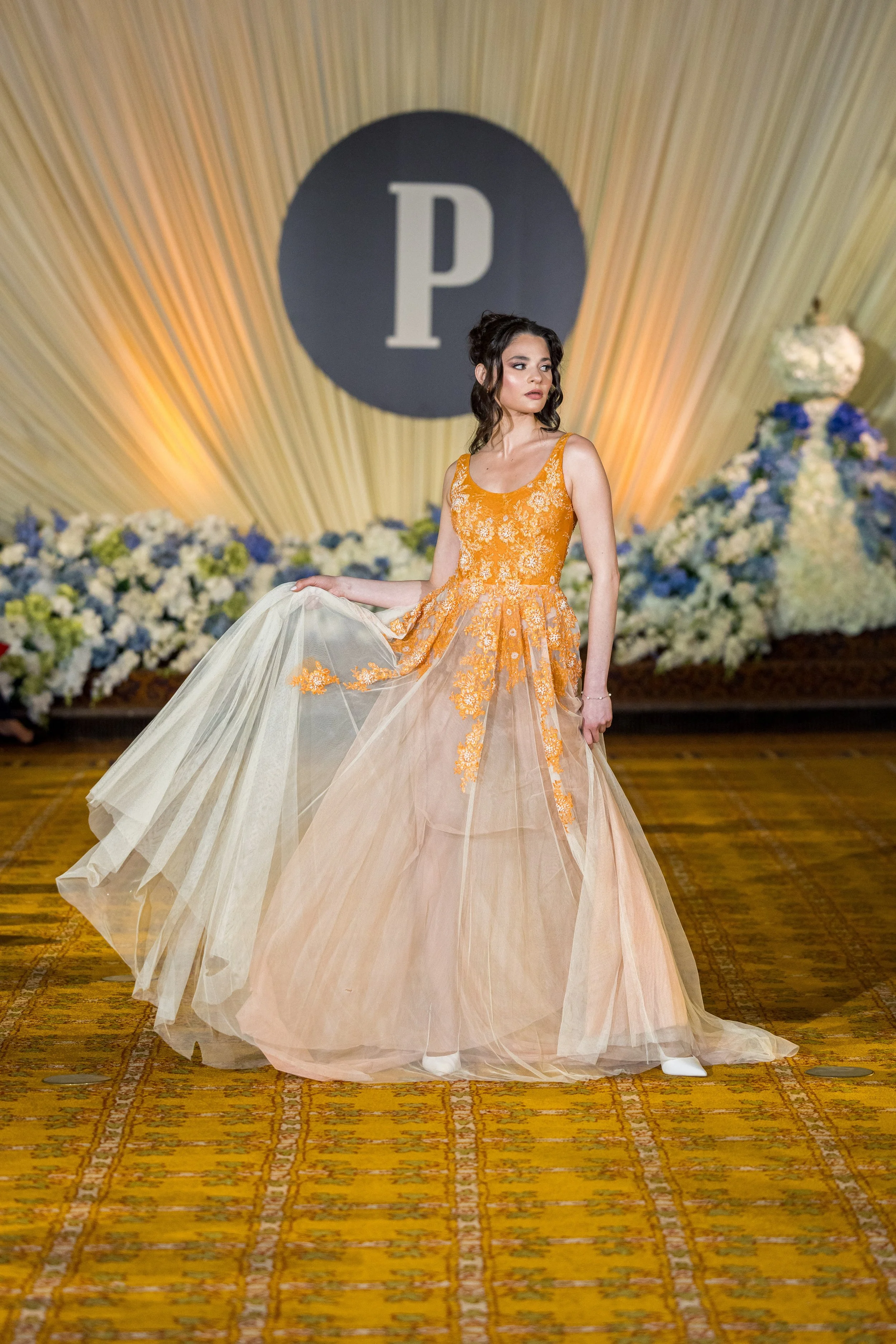 A woman in a yellow and orange floral gown walking on a patterned yellow carpet, with her hand holding up part of her dress. She is standing in front of a backdrop with a large black circle containing a white letter 'P,' and there are floral arrangem