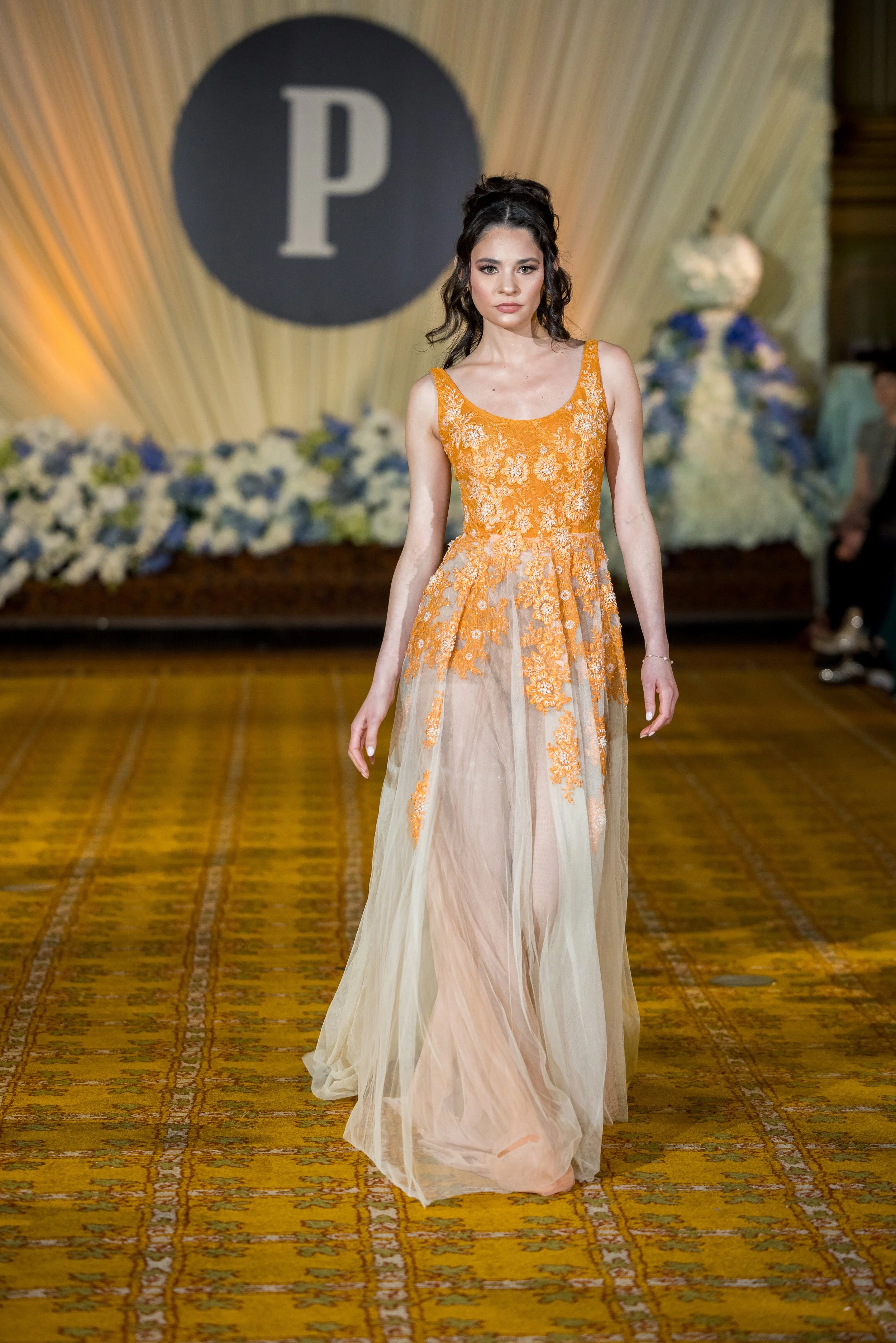 A female model walking on a runway wearing a long, sleeveless orange and beige dress with floral embroidery on the bodice and a sheer flowing skirt. The background features a large black circle with a white letter 'P' and draped fabric with flowers.