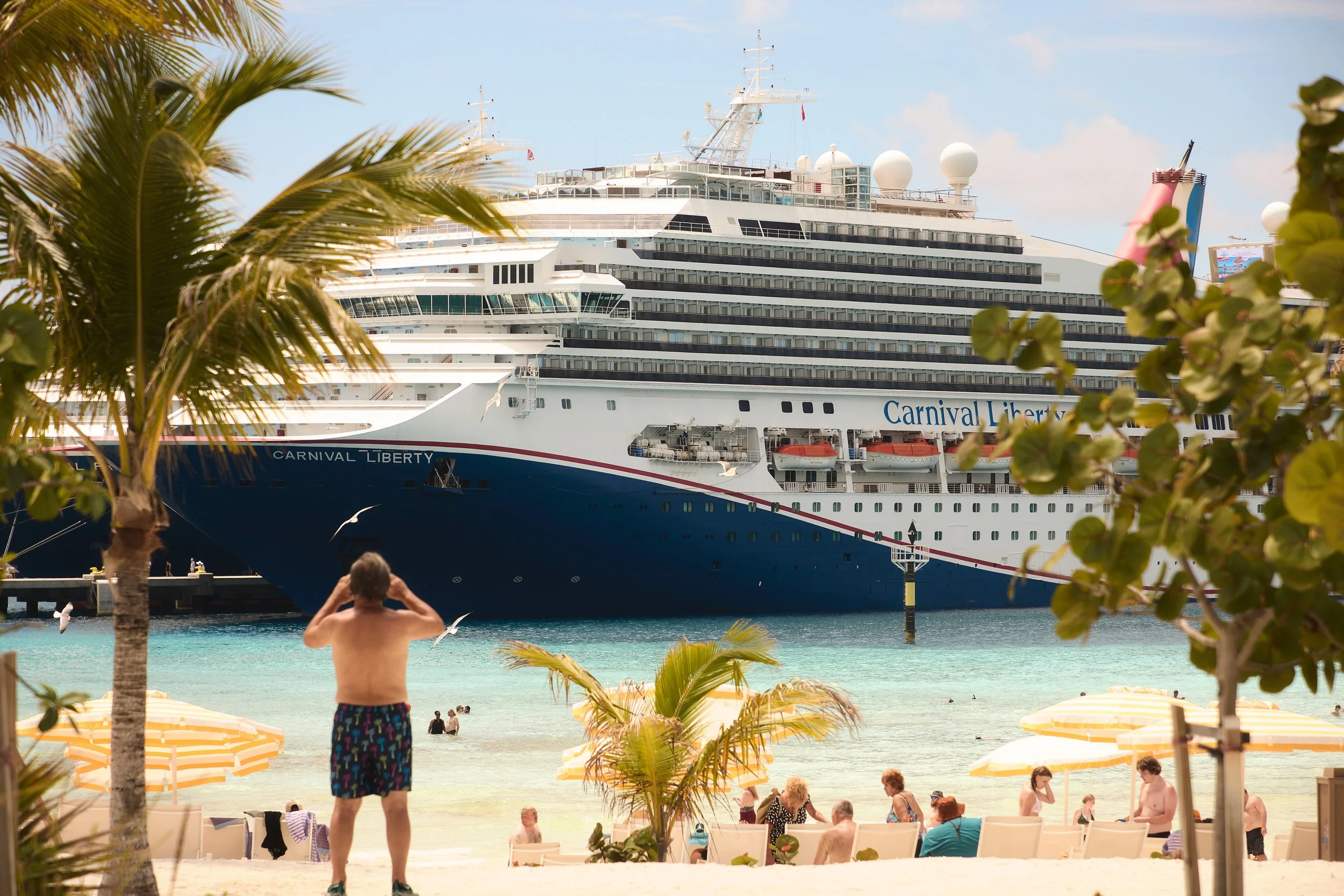 A large cruise ship named Carnival Liberty docked near a beach with people sunbathing and swimming. There are palm trees and yellow umbrellas in the foreground, and a man in colorful shorts stands facing the water. The sky is partly cloudy.