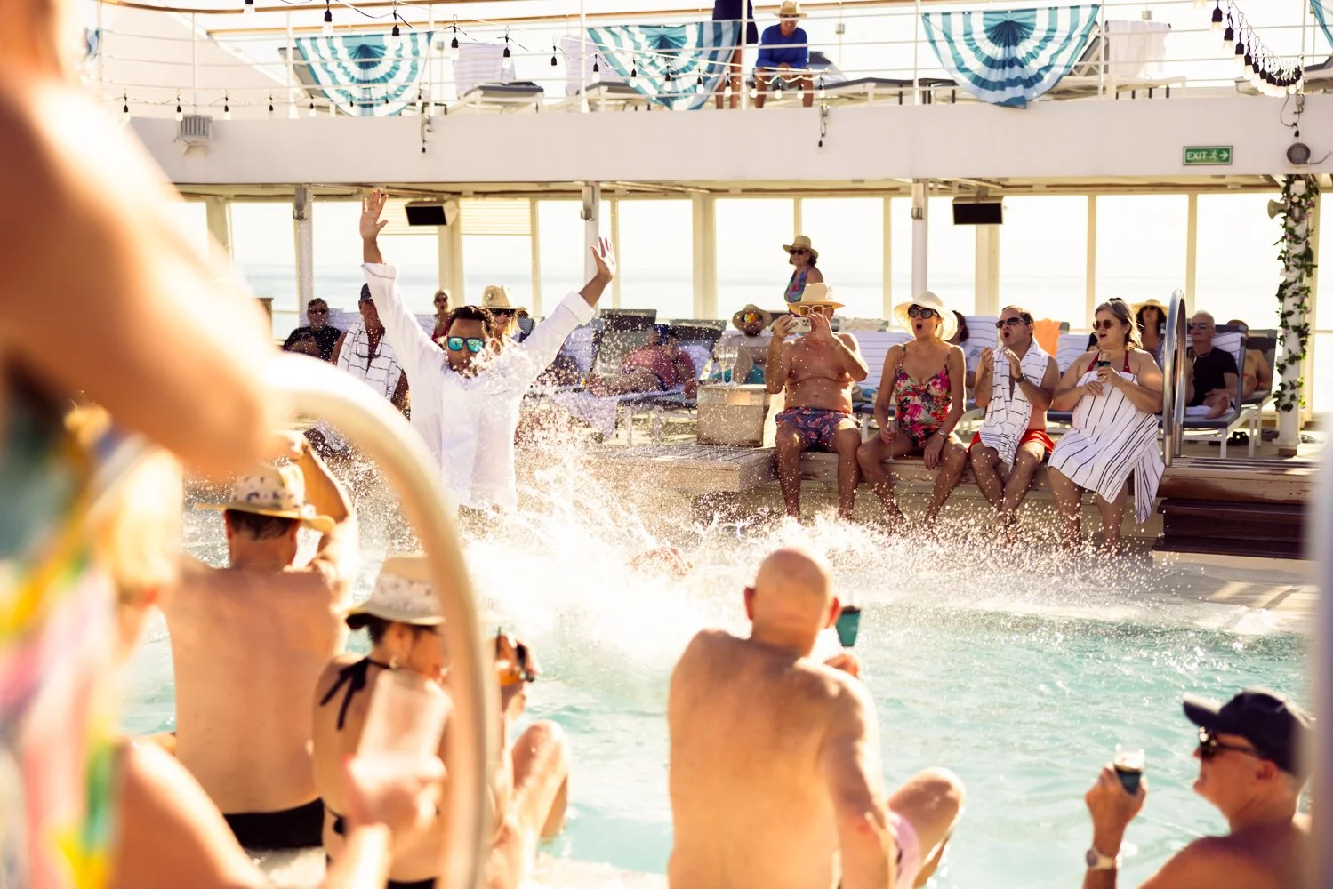 People having fun at a pool party on a cruise ship deck, with some sitting on the edge in swimwear, others taking photos or recording, and a DJ in the background, under decorated canopies and string lights.