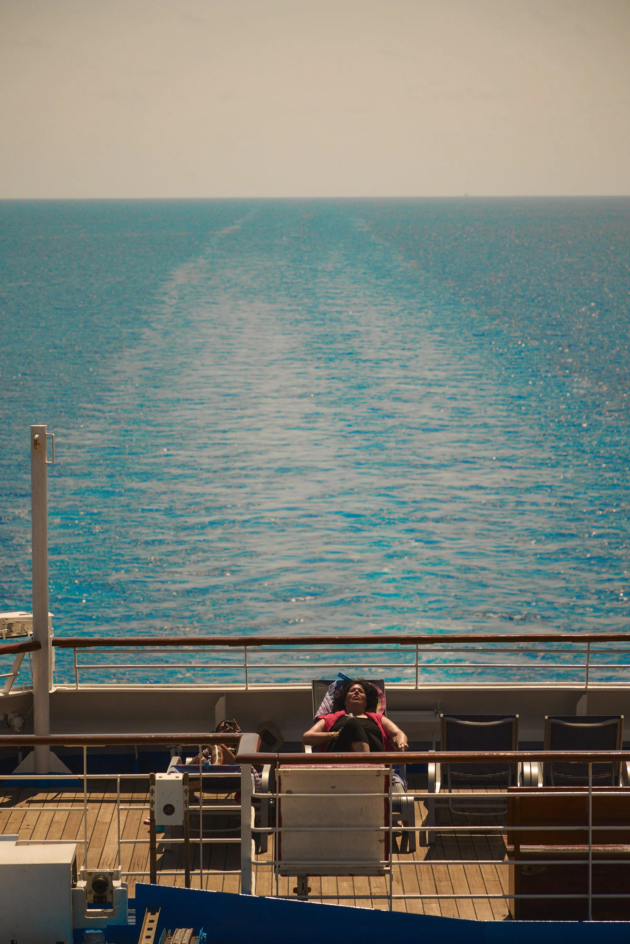 A woman relaxing on a deck chair on a cruise ship, with calm blue ocean and clear sky in the background.