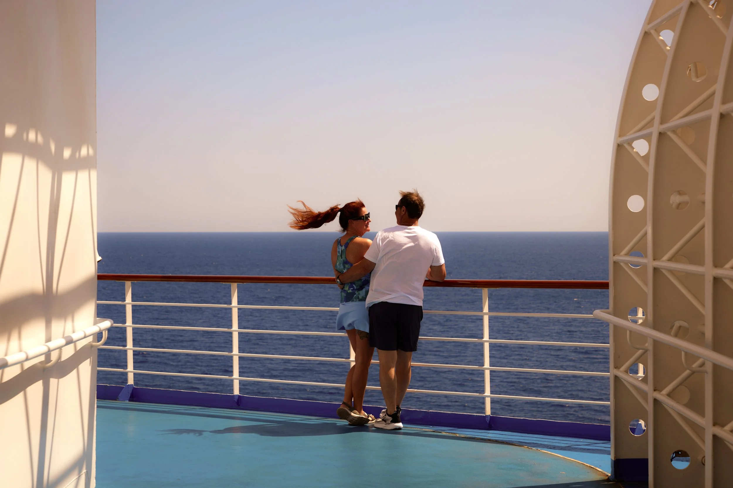 A couple is standing on the deck of a cruise ship, looking out over the ocean, with the woman wearing sunglasses and the man leaning towards her in casual shorts and t-shirt, framed by the ship's structure.