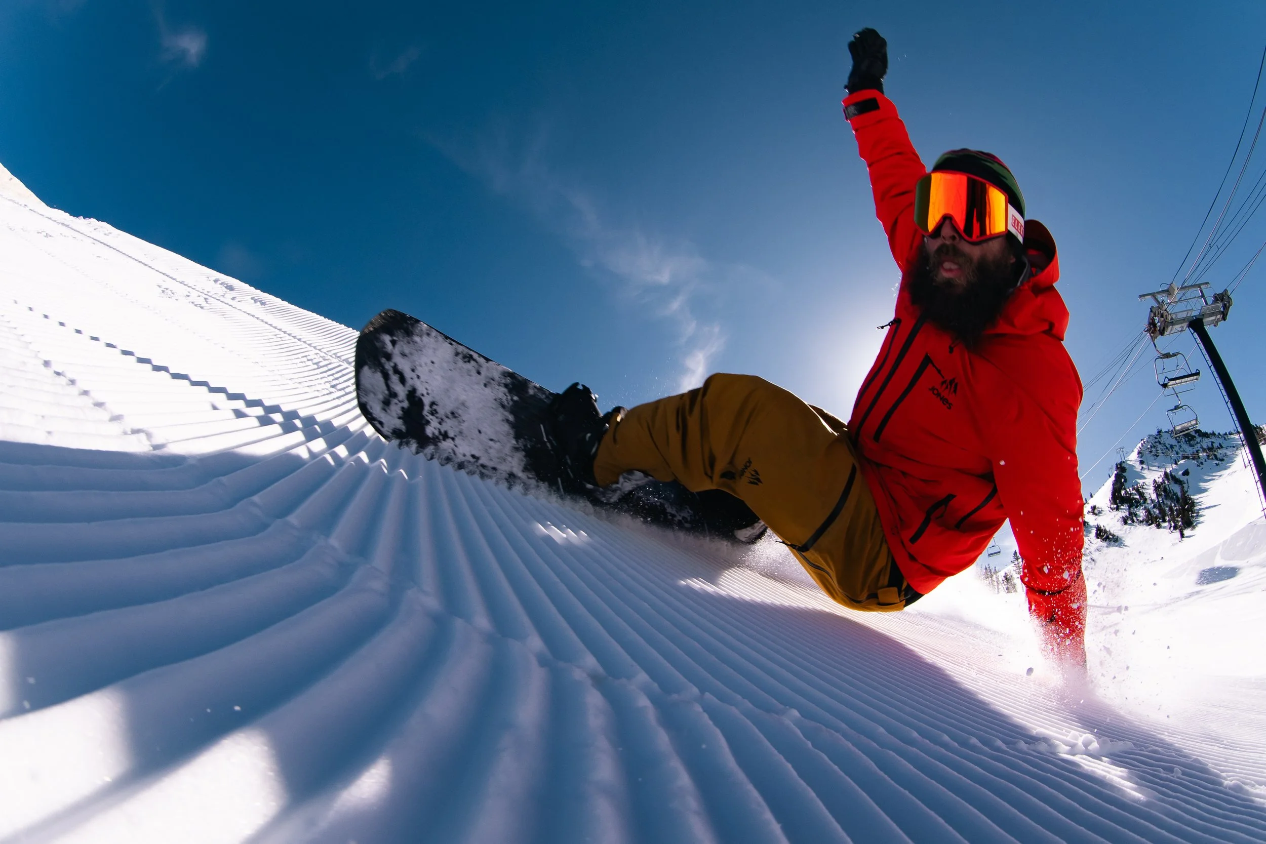 A snowboarder in a red jacket and yellow pants is falling backward on a snow-covered slope with a blue sky and ski lift in the background.