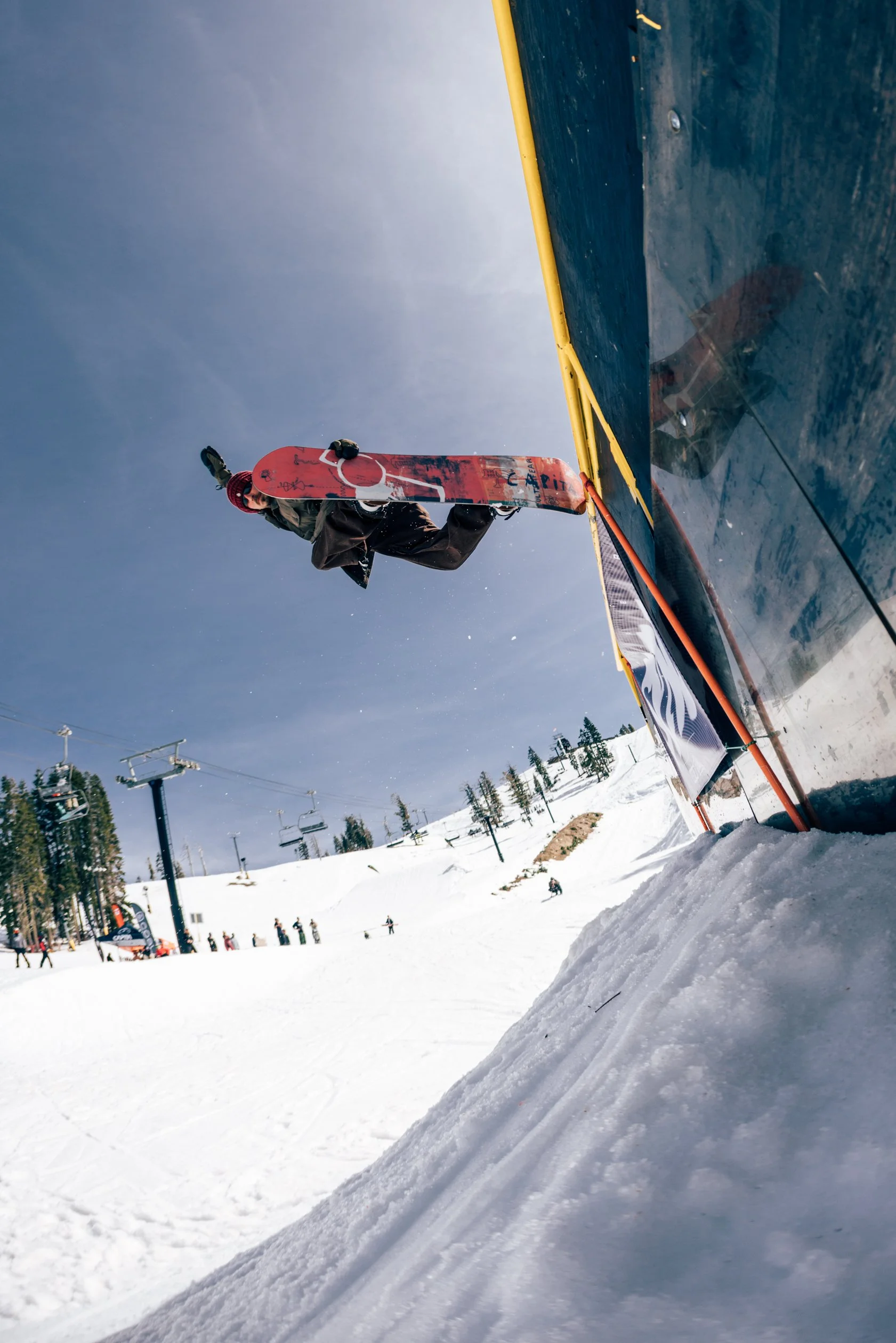 A snowboarder standing on a snow ramp on a ski slope, holding their snowboard, with a ski lift in the background and people skiing in the distance.