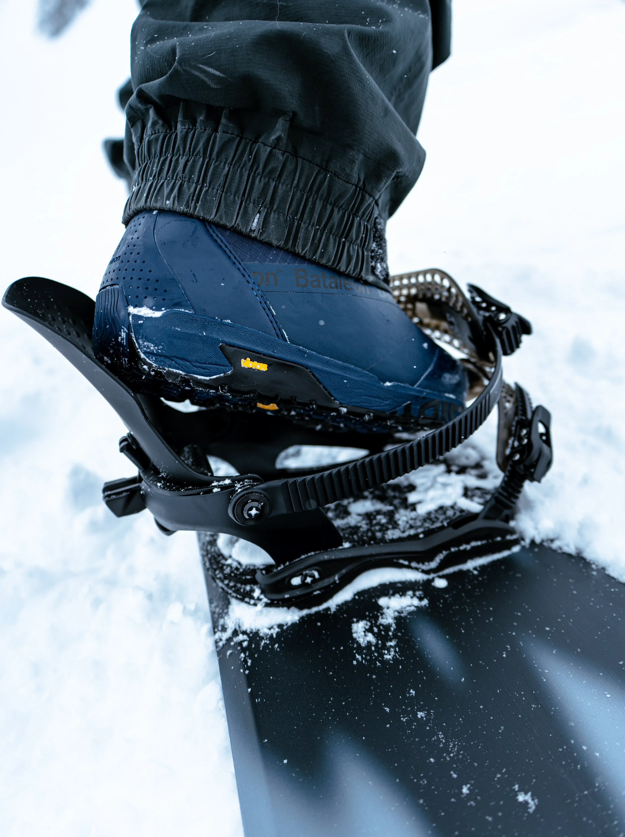 Close-up of a person wearing black snow pants, a black jacket, and blue snow boots with yellow Vibram logo, sitting on a black snow sled in a snowy environment.