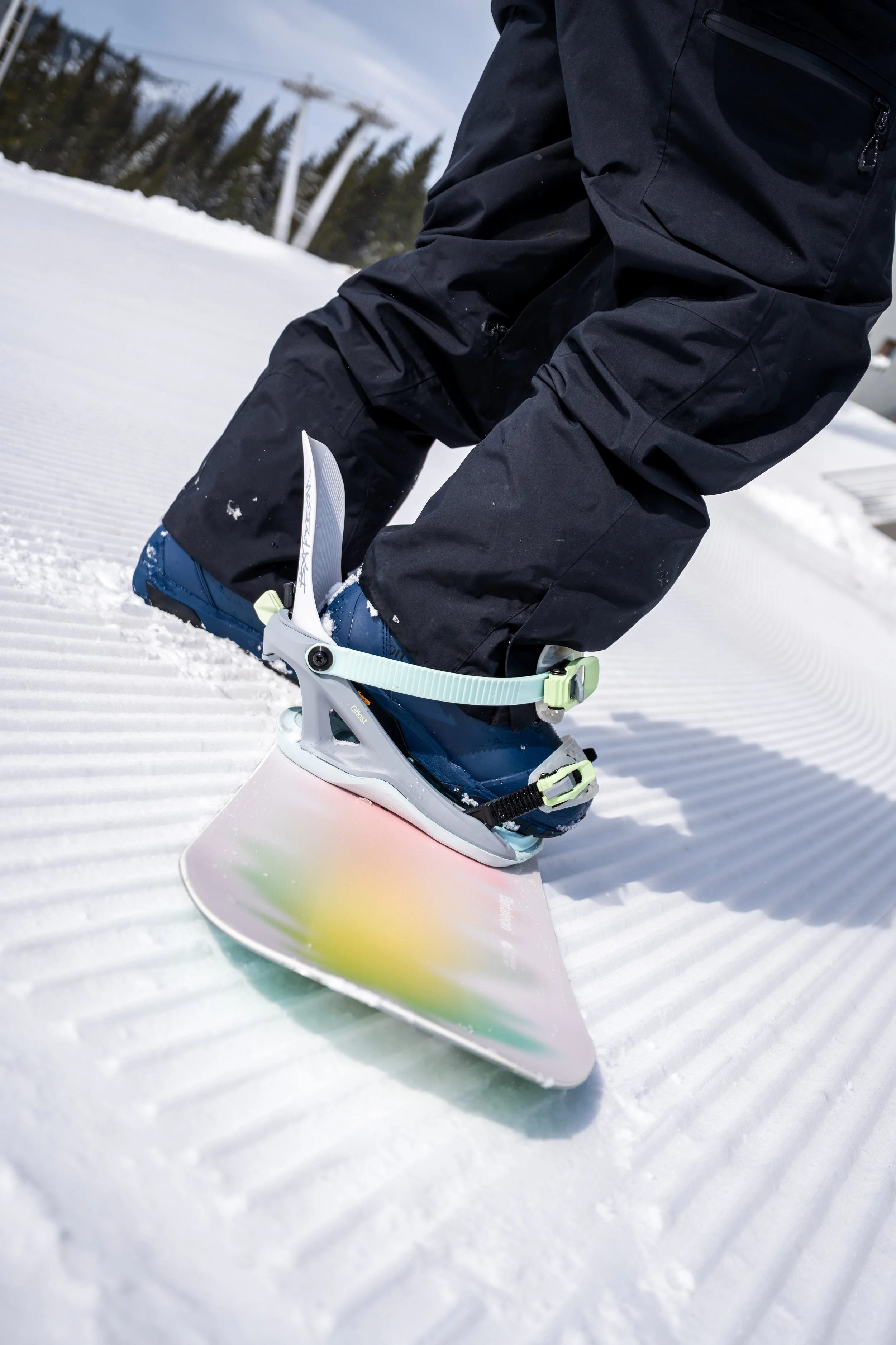 Close-up of a person in black winter gear, wearing blue snowboard boots, tightening bindings on a snowboard on a groomed snow slope.