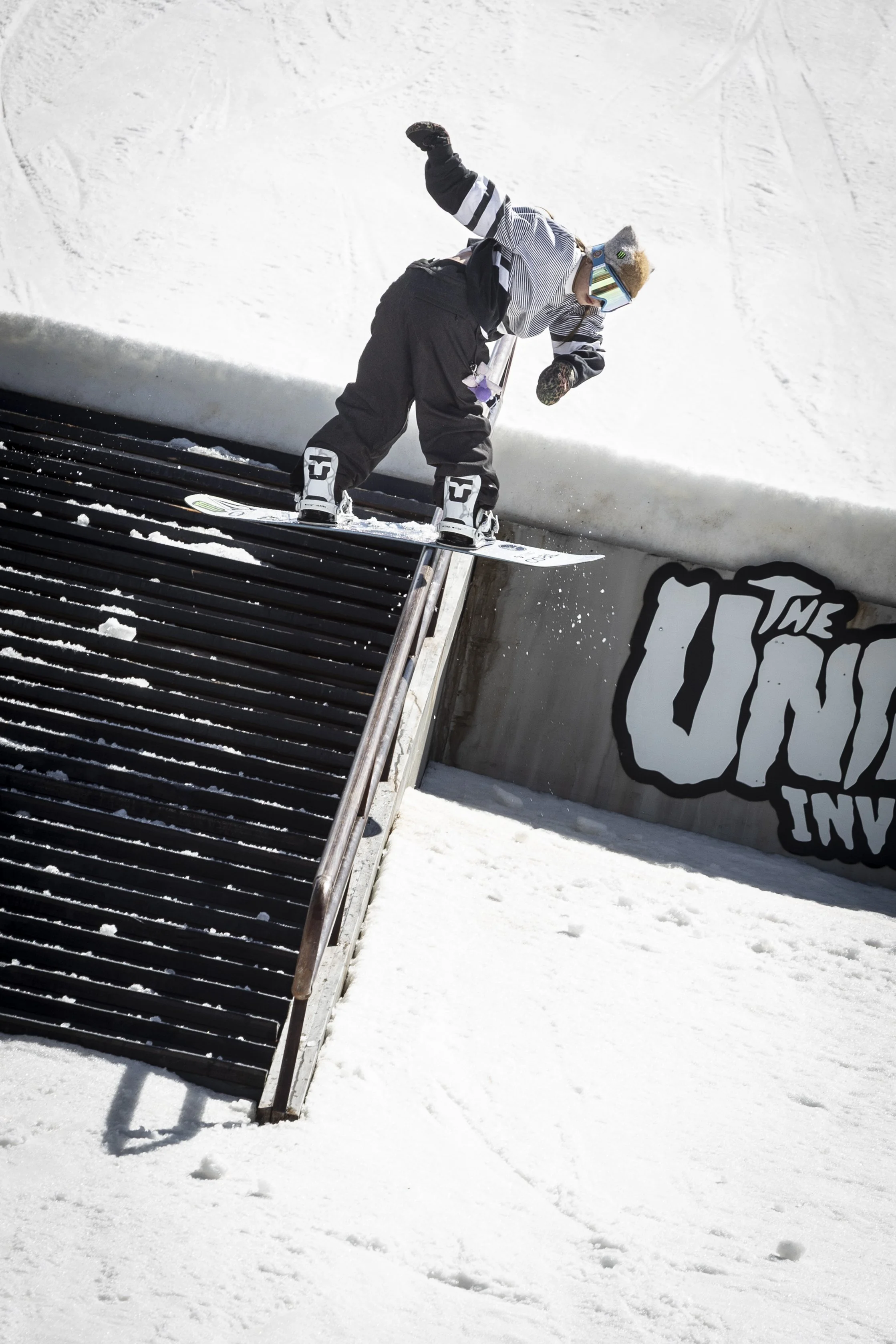 A skateboarder in winter gear riding down a metal rail on a snowy terrain at a snowboarding or skateboarding park, with a graffiti-style wall in the background.