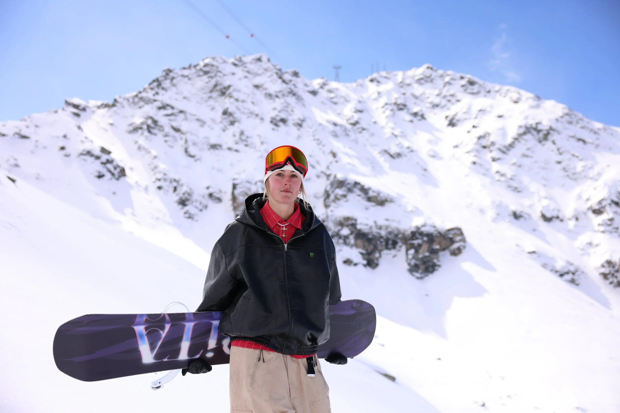 A young person in winter clothing and ski goggles holding a snowboard stands on snow with a mountain and cable car in the background.