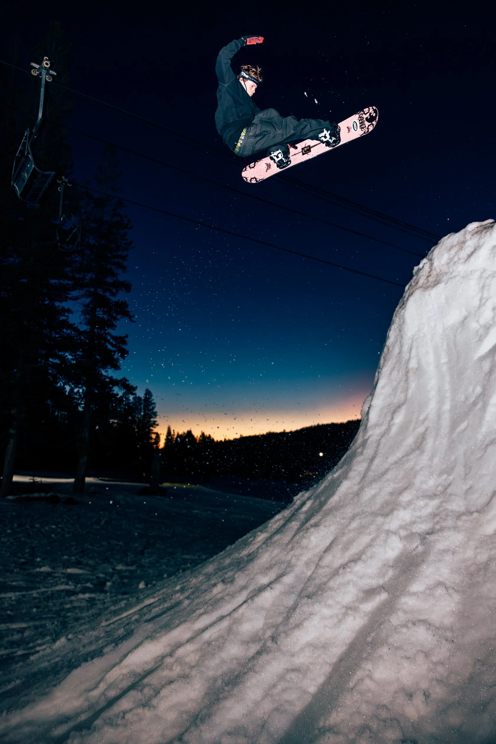 A snowboarder mid-air performing a jump at night, with snow flying around, a snow-covered slope, trees, and a dark sky with stars and a sunset in the background.