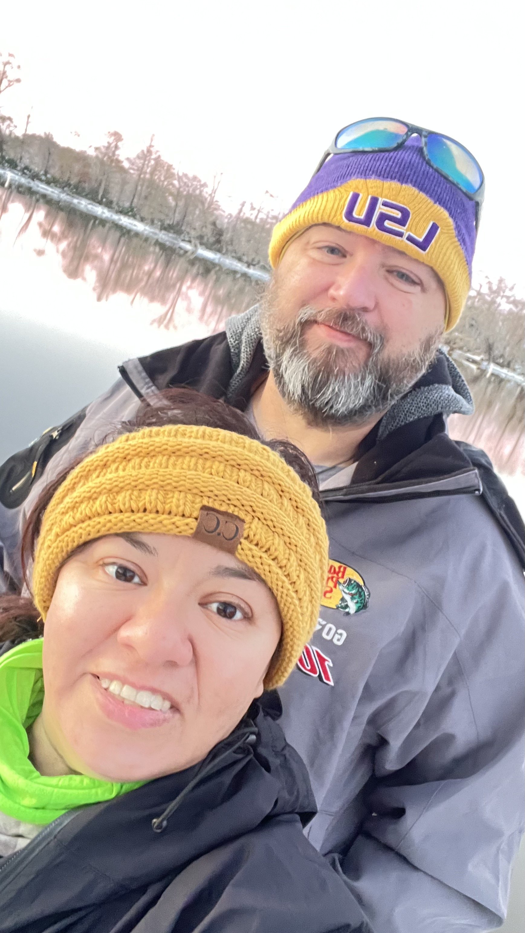Two people wearing winter hats and jackets take a selfie outdoors near a body of water.