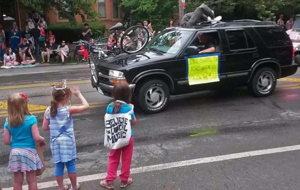 The sign says "I'll share the road when you follow the rules"; there's a crumpled bike embedded in the car, and a dummy dressed as a cyclist hanging through the sunroof. The other dummy is driving. 