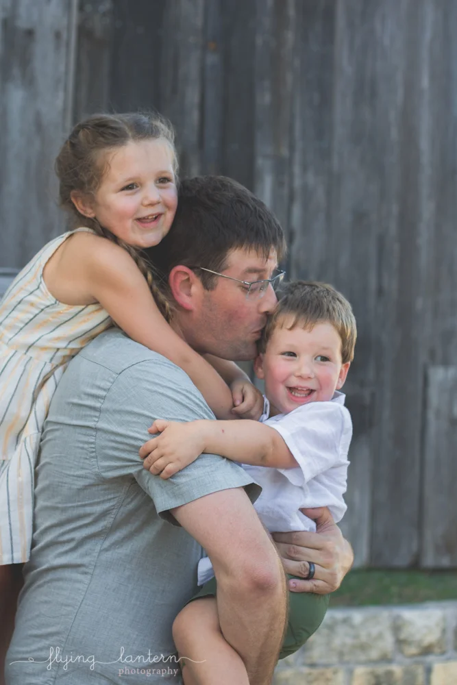father giving daughter piggy back and holding son while giving kiss