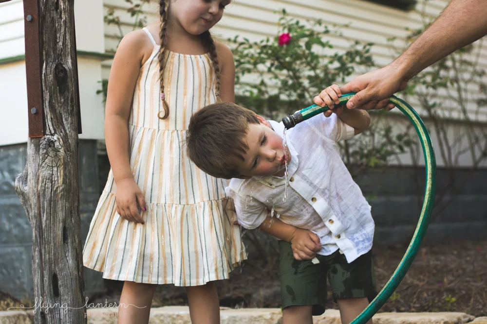 little boy drinking water from hose and sister watching