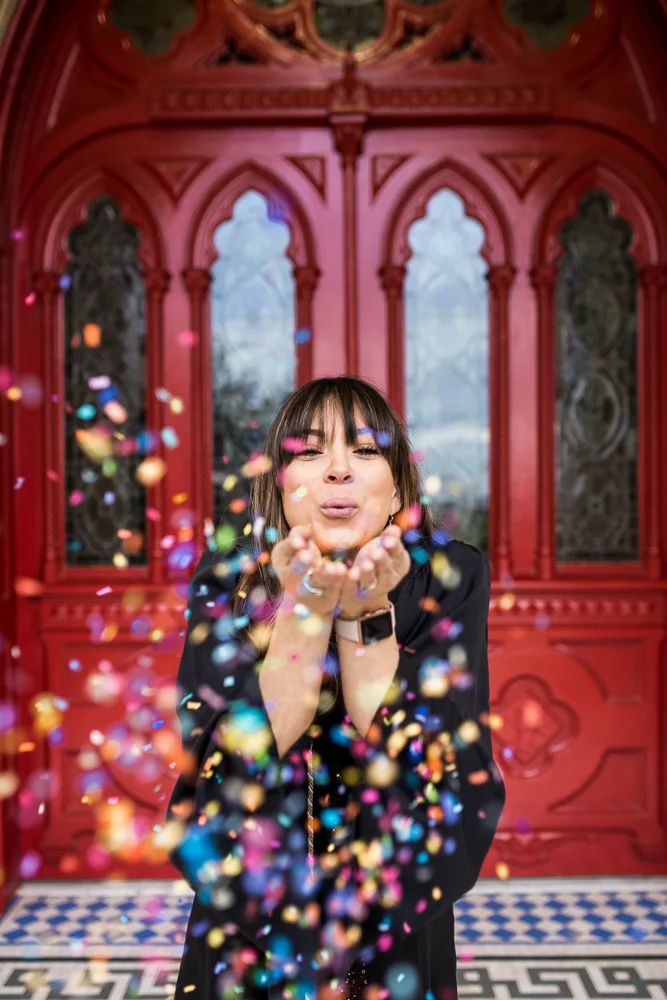 Graduate student at St. Edward’s University in Austin, TX posing at main building in front of red doors blowing confetti. Senior portrait by Erin Reas of Flying Lantern Photography.