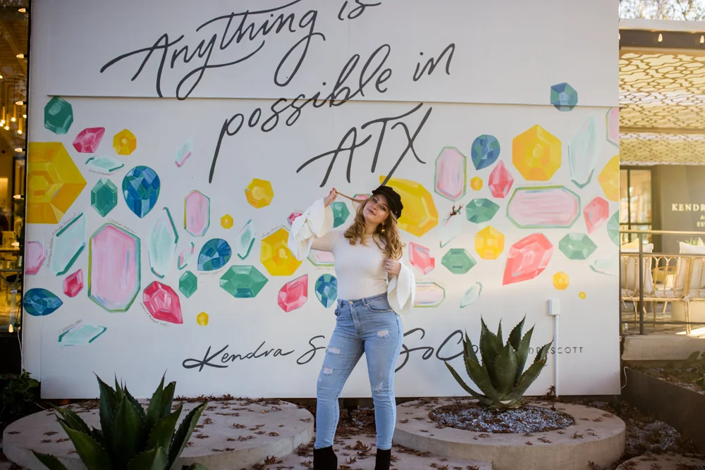 Girl senior portrait outside Kendra Scott wearing white sweater with bell sleeves and black newsboy cap in front of Anything is Possible mural. Photo by Erin Reas photographer in Austin, TX