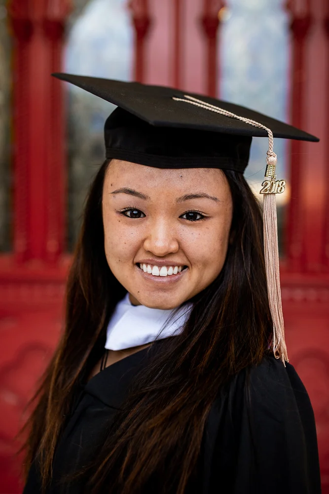 Female college graduate wearing cap and gown standing in front of red doors at St. Edward’s University in Austin, TX. Photo by Erin Reas local senior photographer.