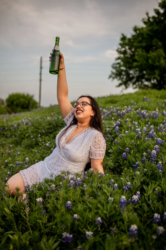 Female college graduate sitting in a field of bluebonnets at St. Edward’s University while drinking a bottle of champagne. Photo by Erin Reas senior photographer in Austin, TX