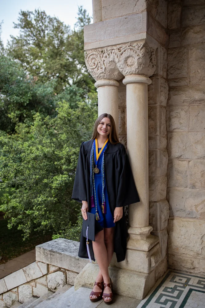 Female college graduate wearing cap and gown standing at the top of St. Edward’s University Main Building in Austin, Texas. Photo by Erin Reas senior photographer