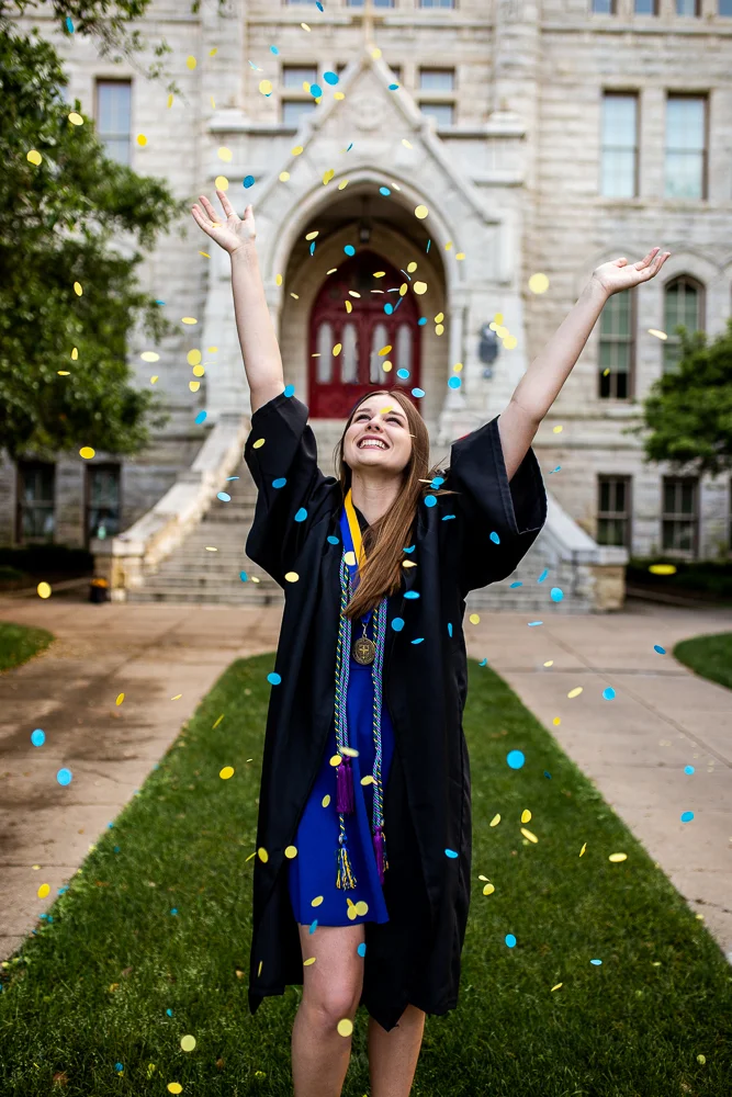 Female college graduate throwing blue and yellow confetti in front of Main Building at St. Edward’s University. Photo by Erin Reas senior photographer in Austin, Texas