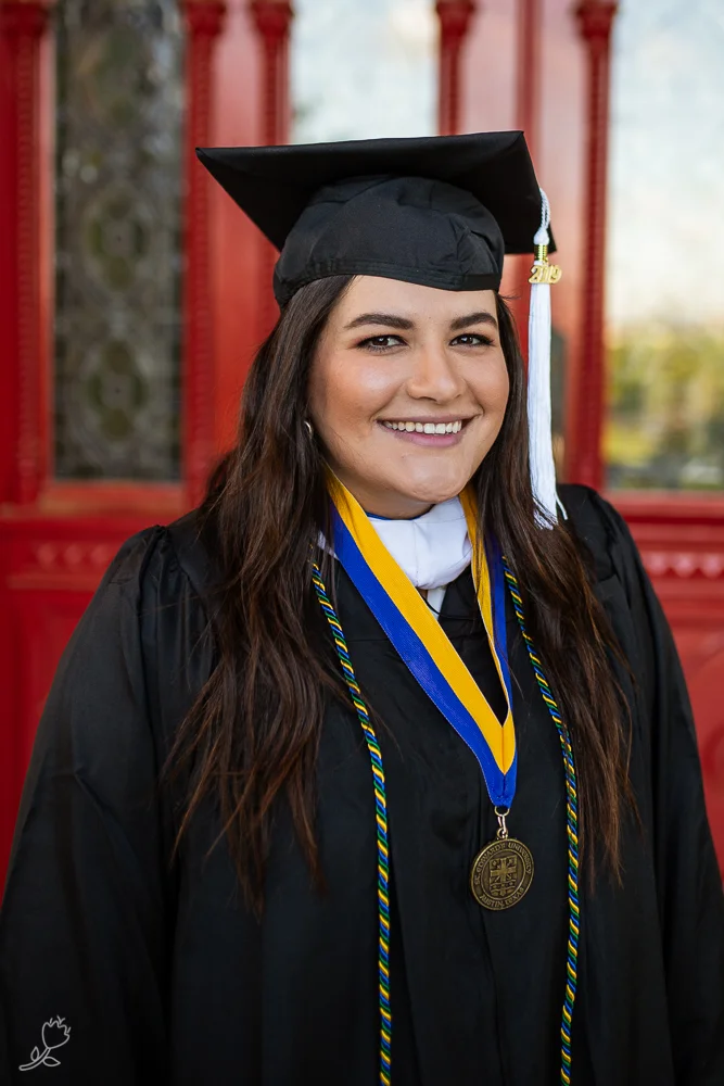 Female college graduate wearing cap and gown standing in front of red doors at St. Edward’s University.