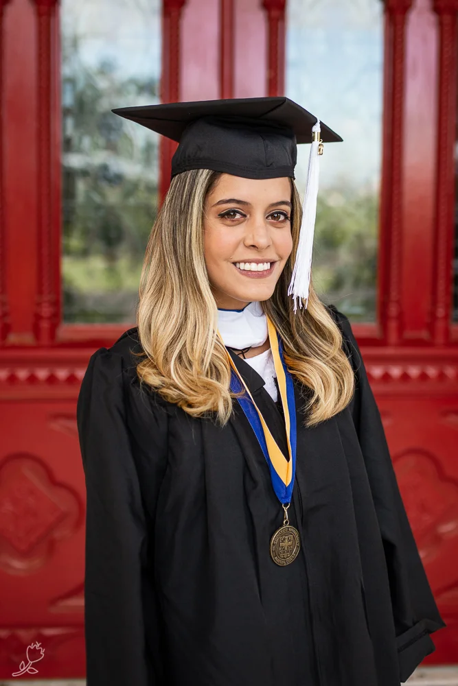 Female college graduate wearing cap and gown standing in front of red doors at St. Edward’s University.