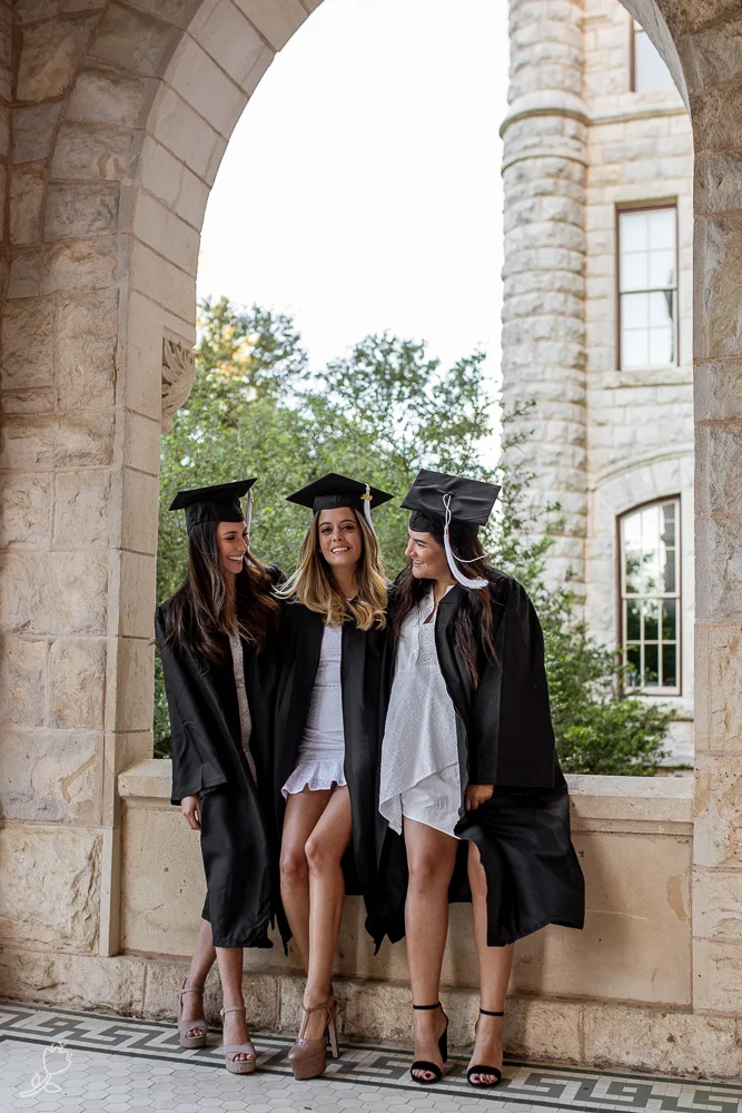 Three best friends in college wearing graduation gowns, caps and white dresses at St. Edward’s Univeristy Main Building