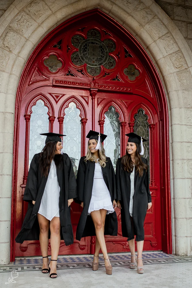 College best friends standing in front of red doors at St. Edward’s University while wearing cap and gown walking away from the doors