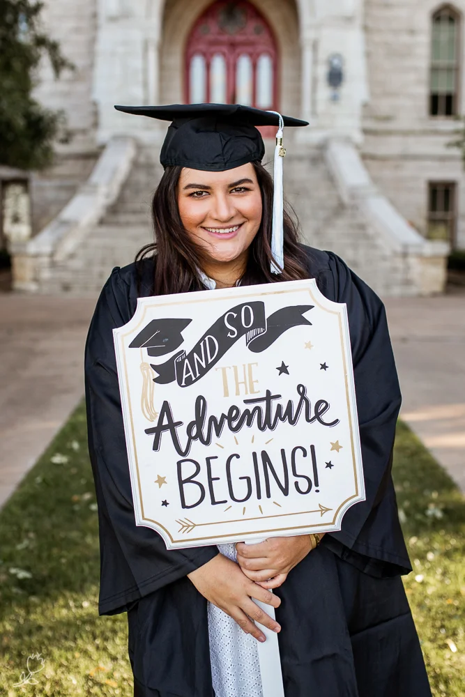 Female college graduate at St. Edward’s University holding graduation sign in front of Main Building. Photo by Erin Reas Austin senior photographer.