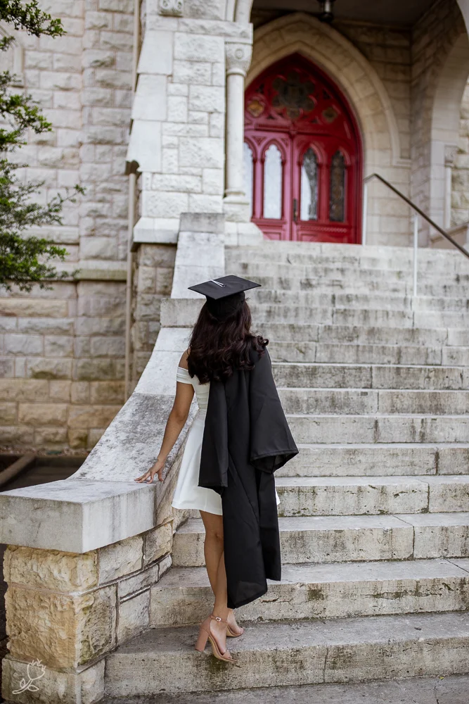 Female college graduate standing at base of Main Building at St. Edward’s University walking up steps while wearing cap and gown over a white dress. Photo by Erin Reas senior photographer
