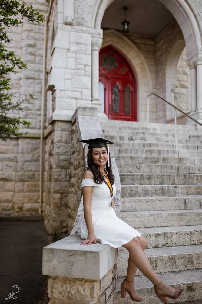 Female college graduate wearing white dress and graduation cap sitting at base of Main Building at St. Edward’s University. Photo by Erin Reas senior photographer in Austin, TX