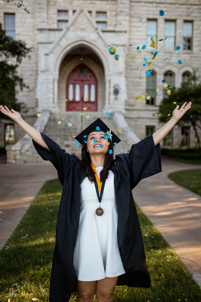 St. Edward’s University college graduate throwing blue and yellow confetti in front of Main Building. Photo by Erin Reas senior photographer in Austin, TX
