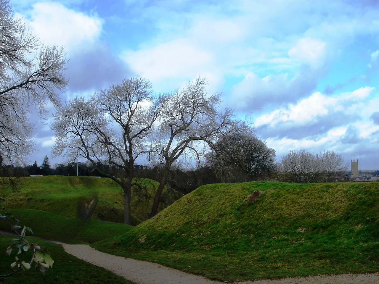A scenic park with leafless trees, grassy mounds, a winding dirt path, and a cloudy blue sky, with a tower visible in the distance on the right side.