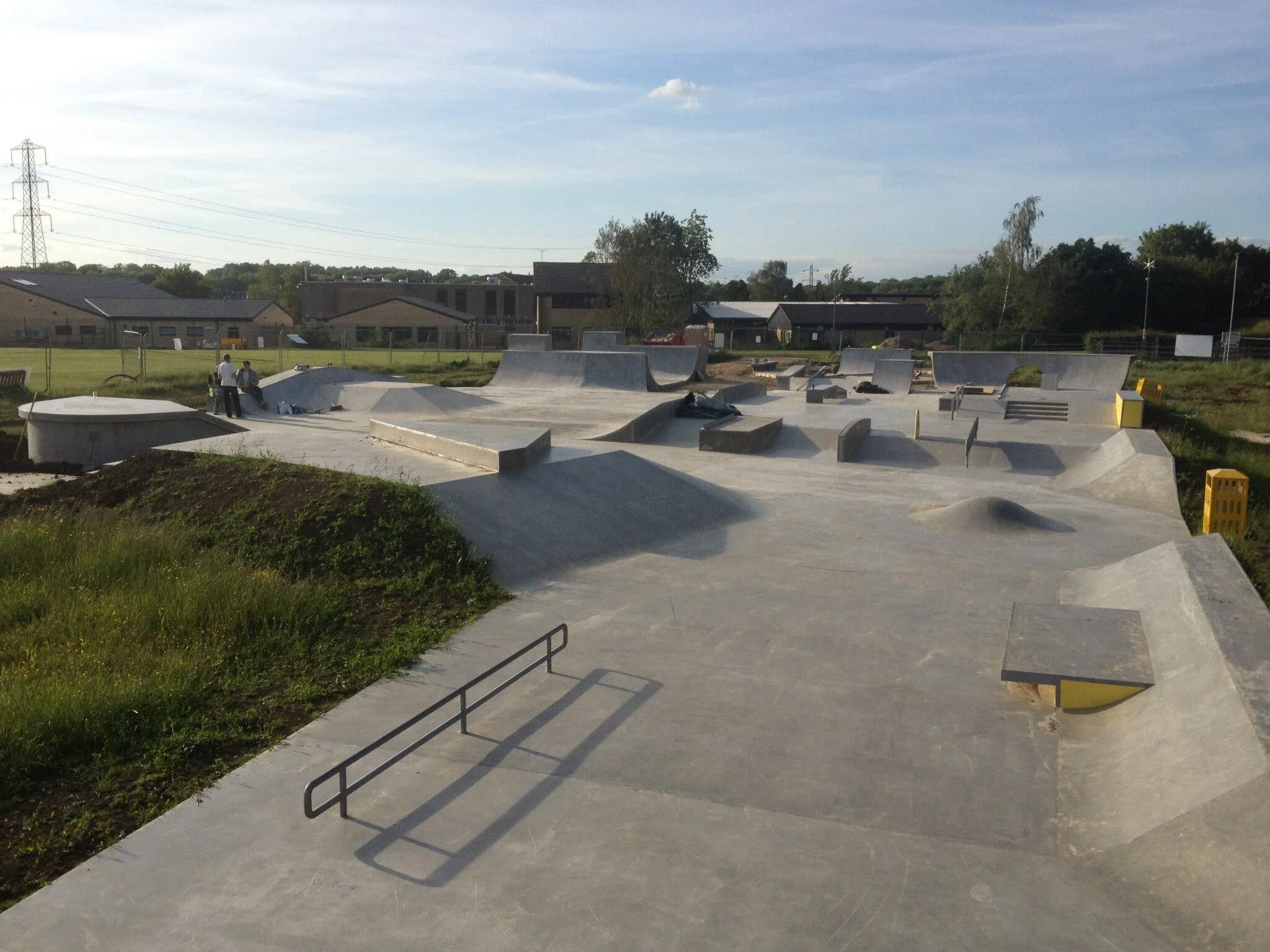 Concrete skatepark with various ramps, rails, and ledges under a partly cloudy sky, with buildings and trees in the background.