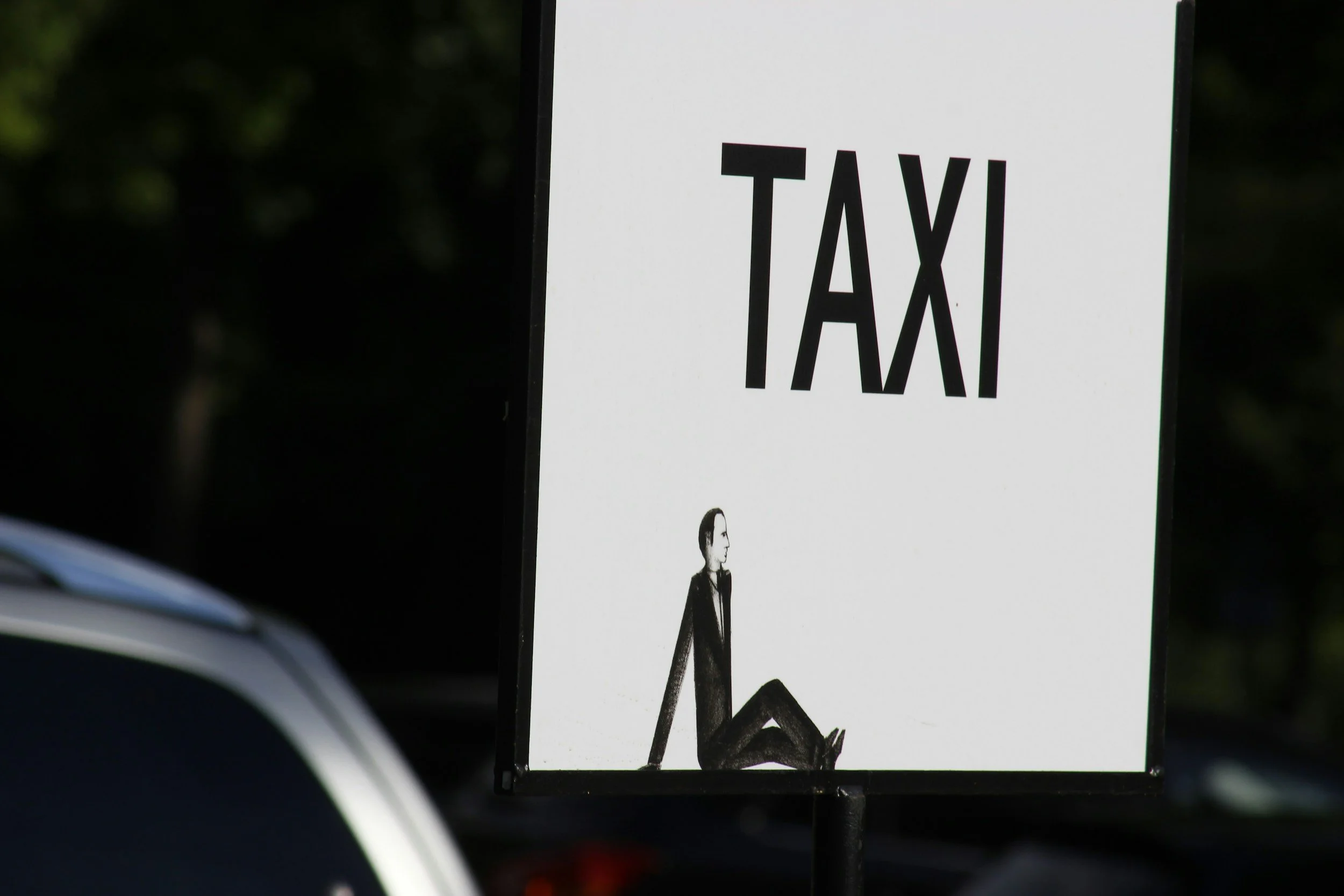 A parking sign with the word 'TAXI' in large black letters and a black-and-white illustration of a sitting woman on a white background.