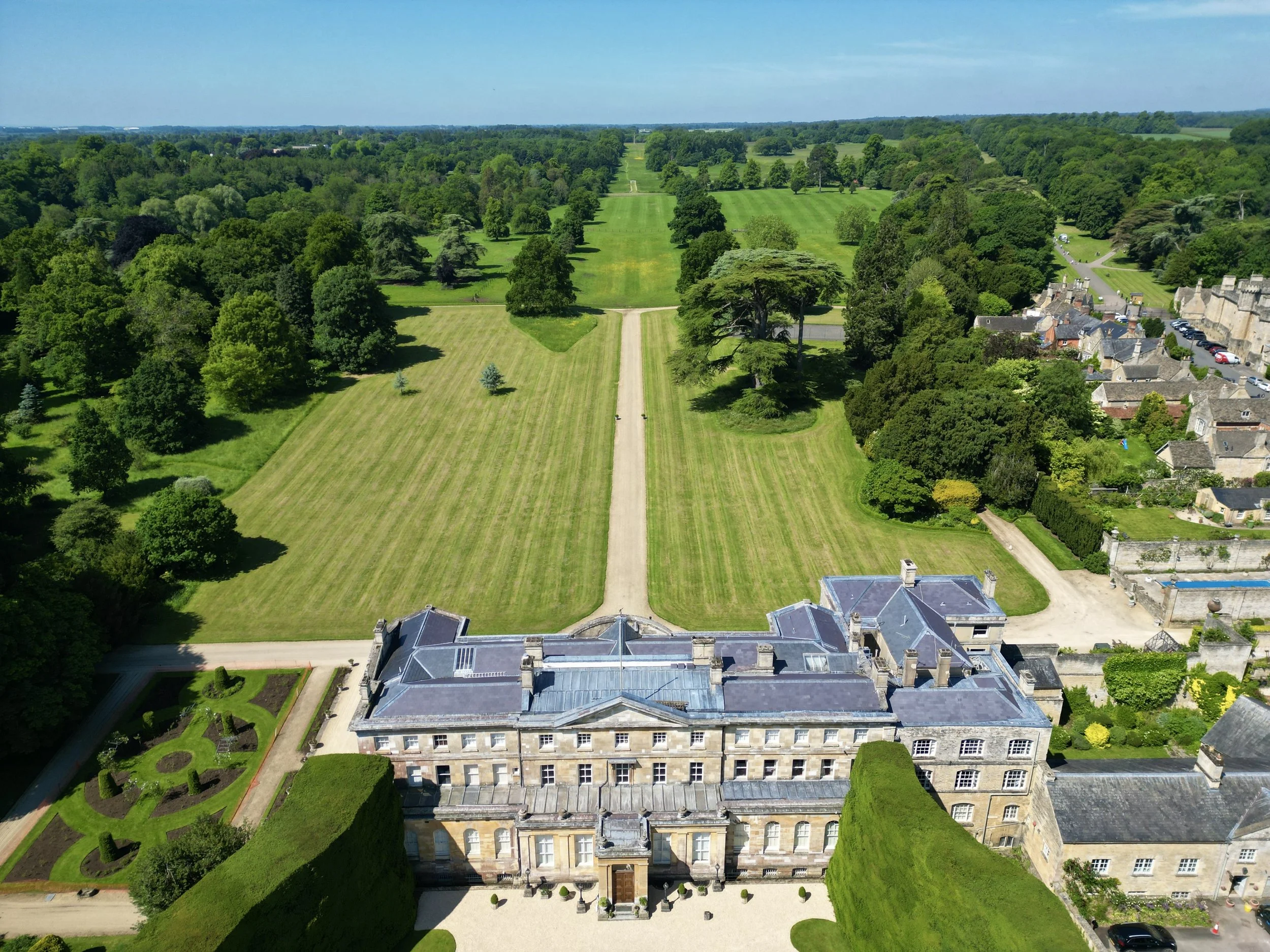 Aerial view of a historic chateau surrounded by well-manicured gardens, expansive green lawns, and trees, with a small town on the right and a vast countryside in the background.