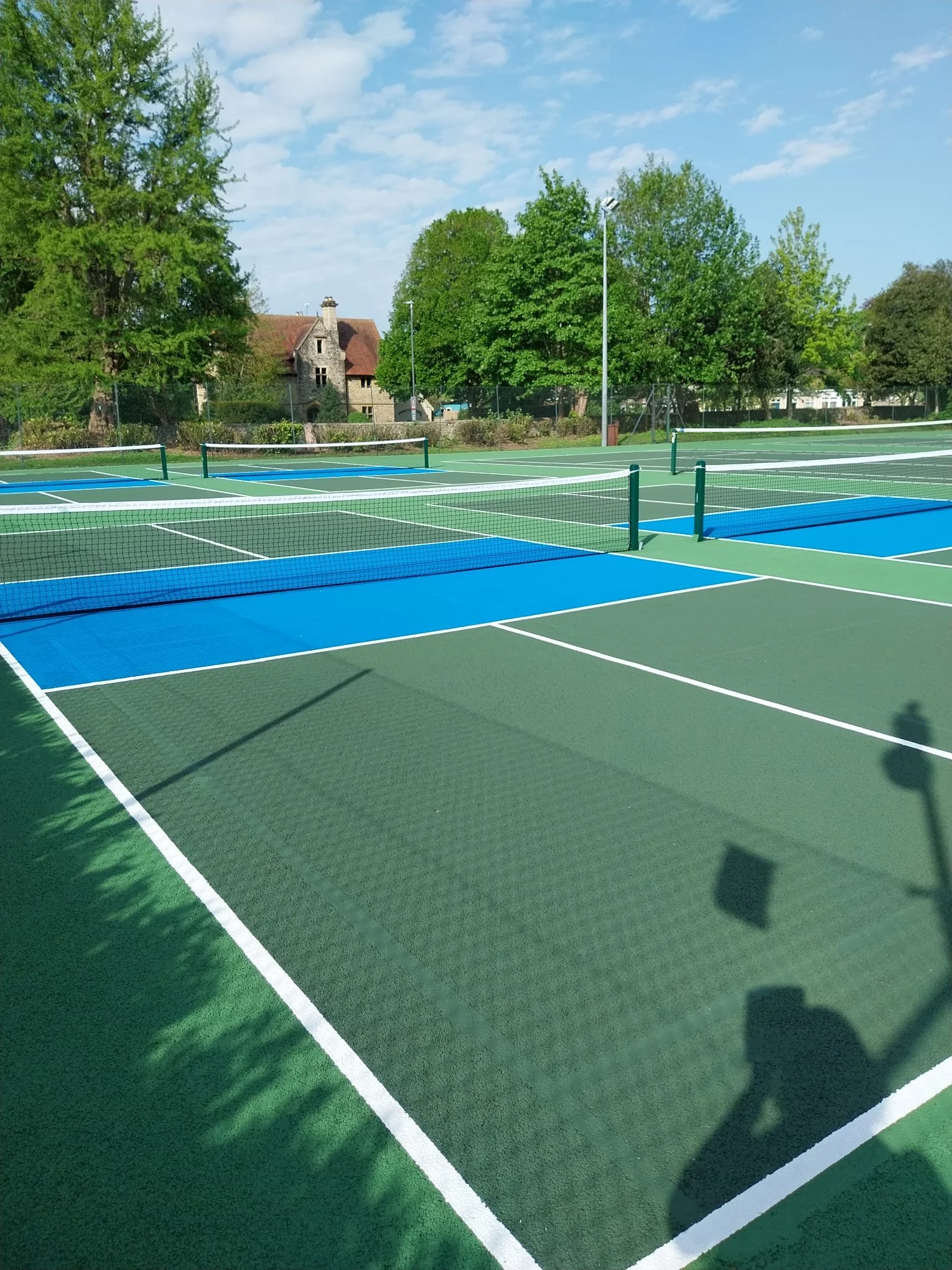 Empty outdoor pickleball courts with blue and green surfaces, surrounded by trees and a cloudy sky.