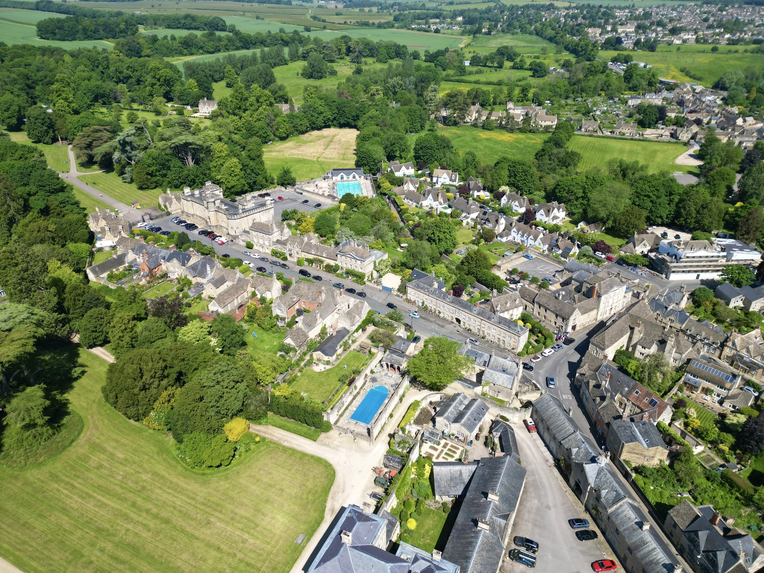 Aerial view of a small town surrounded by lush green fields and trees, with a castle, swimming pool, and various residential buildings.