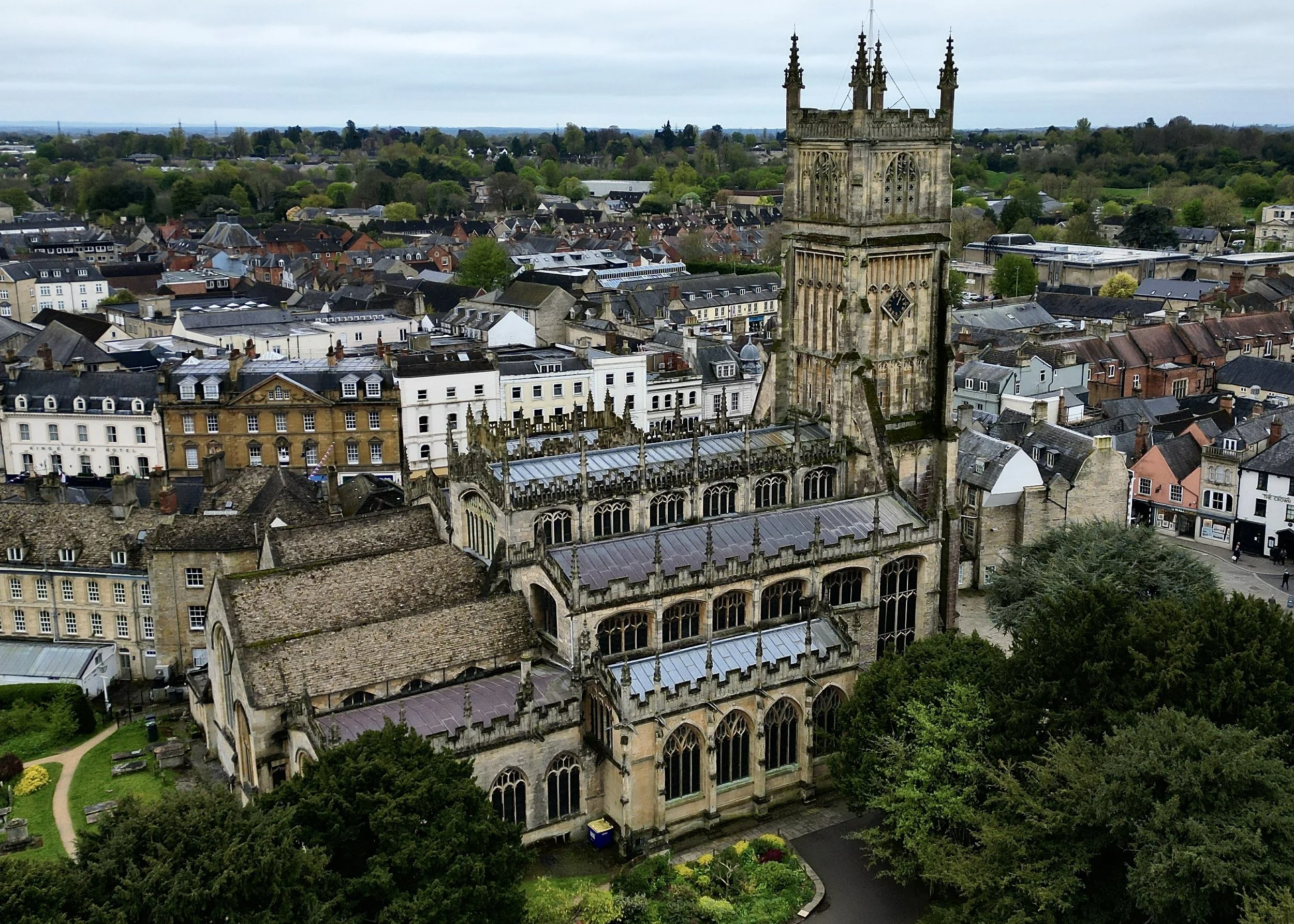 Aerial view of a historic stone church with a tall tower, surrounded by trees and a cityscape with various buildings and rooftops.