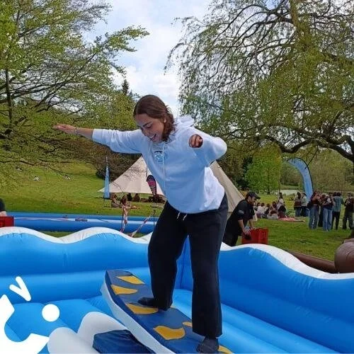 A woman balancing on an inflatable surfing simulator outdoors, with a park and other people in the background.
