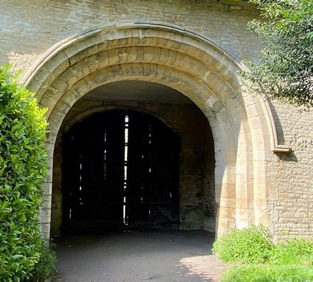 An arched stone gateway with a dark wooden door, surrounded by greenery, possibly part of an old castle or historical building.